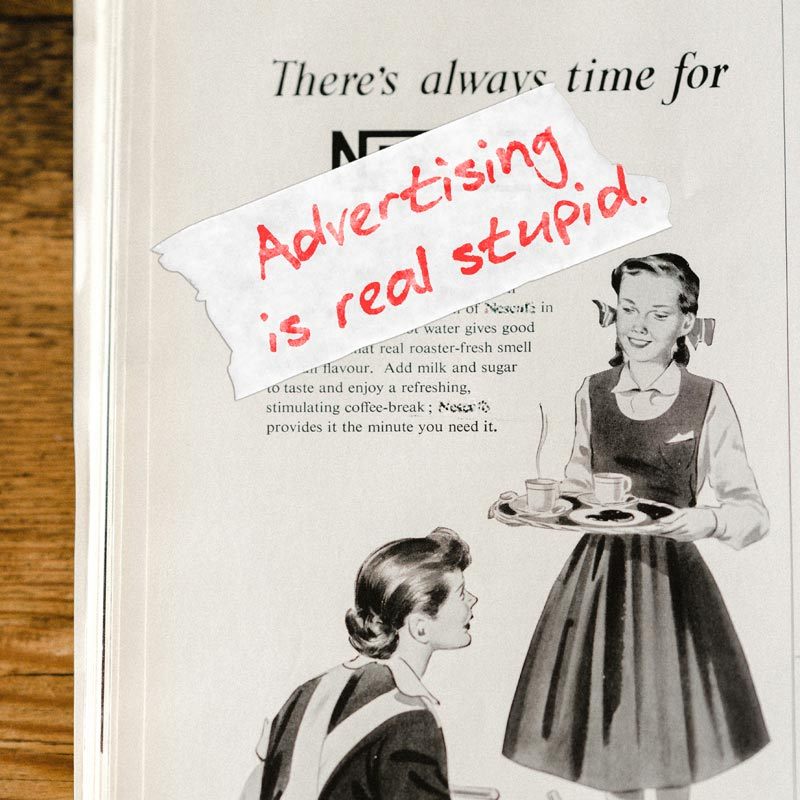 Image: A magazine laying on a wood table, open to an old black-and-white ad, with a piece of white tape stuck across the ad saying, 'Advertising is real stupid.'